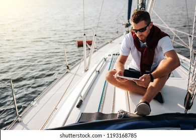 Serious And Peaceful Guy Sits On Board Of Yacht. He Holds And Looks At Tablet. Young Man Is Calm. He Wears White Sirt And Dark Sweater With Shorts.