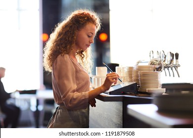 Serious Pensive Attractive Curly-haired Waitress Standing At Bar Counter And Using Pencil While Adding Order In Restaurant POS