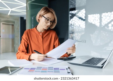 Serious and pensive business woman behind paper work inside office, female financier worker thinks about contracts and reports with charts and graphs, blonde successful woman uses laptop at work. - Shutterstock ID 2321363417