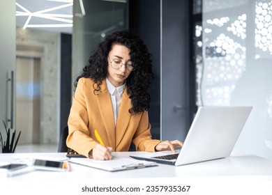 Serious and pensive business woman behind paper work inside office, female financier worker thinks about contracts and reports with charts and graphs, hispanic successful woman uses laptop at work