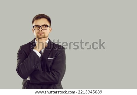 Serious pensive businessman looks at content on copy space which arouses suspicion and doubt. Young man in suit and glasses holding his chin looks to side on gray background. Isolated. Web banner.