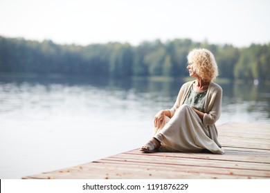Serious pensive curly-haired mature lady in long skirt sitting on pier and contemplating tranquil nature around - Shutterstock ID 1191876229