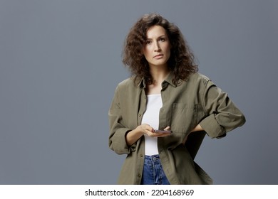 Serious Pensive Doubting Curly Beautiful Woman In Casual Khaki Shirt Can Not Make Decision About Huge Buy Using Phone Posing Isolated On Gray Background. Social Media Influencer Concept. Copy Space