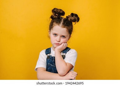 Serious Pensive Little Kid Girl Looking Unhappy At Camera, Tired Upset Preschool Child With Thoughtful Face Holding Hand Under Chin, Melancholic Or Bored, Isolated Over Yellow Studio Background