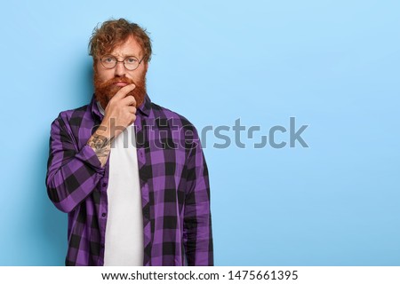 Serious pensive man scowls eyebrows and touches chin, being deep in thoughts, thinks over future plans, wears optical glasses and plaid purple shirt stands indoor against blue wall. Facial expressions