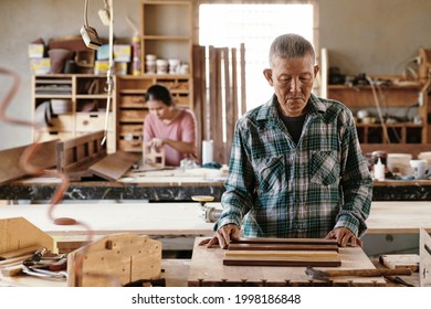 Serious Pensive Senior Carpenter Looking At Pieces Of Walnut And Maple Wood That He Wants To Fuse Together For Making Edge Grain Cutting Board