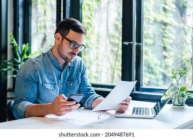 Serious pensive thoughtful focused young casual entrepreneur small business owner accountant bookkeeper in office looking at and working with laptop and income tax return papers and documents
