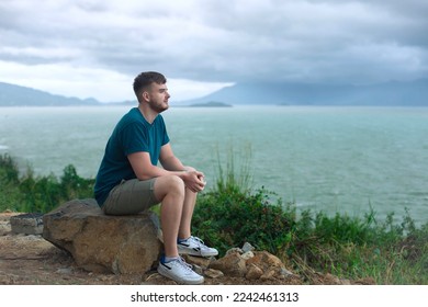Serious pensive thoughtful handsome happy guy, young man enjoying sea, ocean view, landscape, looking into distance at summer day in tropical country at storm with cloudy sky, breathing deep fresh air