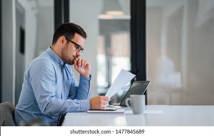 Serious pensive thoughtful young businessman or entrepreneur in modern contemporary office looking at and working with laptop and paper documents making serious and important business decision - Shutterstock ID 1477845689
