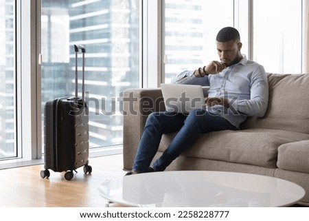 Serious pensive travelling freelance business man using laptop on sofa, working from hotel room, sitting on couch at suitcase, touching chin, thinking over project tasks, looking at monitor