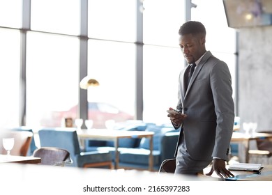 Serious Pensive Young Black Businessman In Stylish Suit Standing In Restaurant And Checking Sms On Phone While Waiting For Business Partner