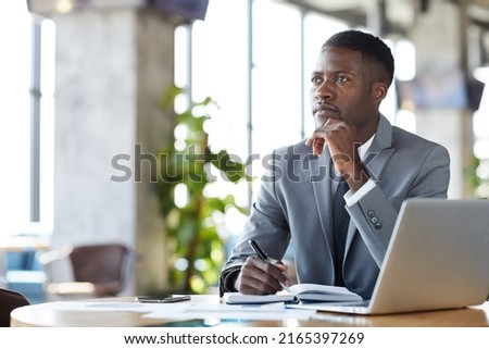 Serious pensive young black manager in formal suit sitting at table and looking into distance while making notes in diary