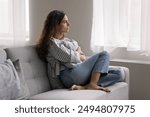 Serious pensive young woman thinking on problem, hugs cushion seated on couch at home, deep on sad thoughts, staring out window, looks depressed, frustrated, misses her beloved, feels lonely. Solitude