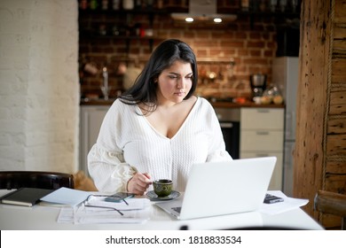 Serious plus size young businesswoman in white blouse working distantly from home office checking email, doing paperwork, sitting at kitchen table with laptop pc, documents and cup of coffee