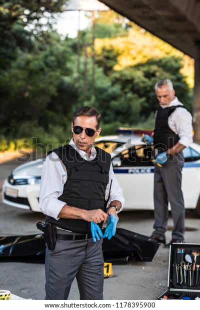 serious policeman in sunglasses putting on latex gloves\
while his colleague standing behind at crime scene with corpse in\
body bag 