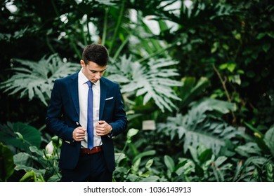 Serious Portrait Handsome Groom In A Wedding Suit And Tie Is Standing On The Background Of Greenery. Man Stands In The Botanical Garden Full Of Greenery.