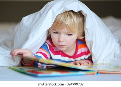 Serious Preschooler Girl Reading Book In Bed. Cute Kid Lying Under The Blanket. Early Learning And Children Development Concept.