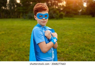 Serious Preteen Boy With Ginger Hair In Blue Superhero Costume And Mask, Looking At Camera While Standing On Green Grassy Meadow With Crossed Hands At Sunset