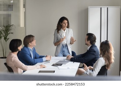 Serious pretty millennial project leader woman talking to employees on meeting, standing at table, explaining tasks, presenting strategy, plan. Female boss mentoring team - Shutterstock ID 2258472153