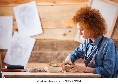 Serious Redhead Young Woman Potter Sitting And Creating Dishes With Clay By Hands