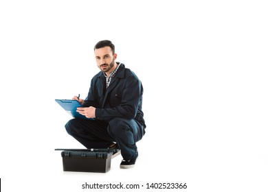 Serious Repairman Writing On Clipboard While Sitting Near Toolbox And Looking At Camera Isolated On White