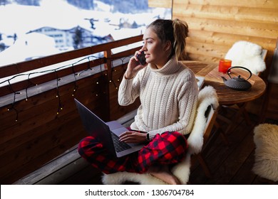 Serious and responsible girl talking on the phone about her work. Laptop lying on her knees, she working remote. Wooden coffee table with teapot and cups. Soft natural sheep fur on the chair.