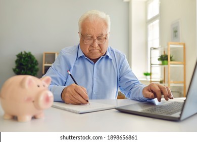 Serious Retired Senior Man In Glasses Using Laptop Computer And Writing In Notebook, Studying Pension Plan, Managing His Budget, Calculating Personal Expenses Or Monthly Annuity Payment