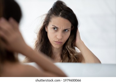 Serious sad millennial girl concerned about dry bad damaged long hair condition, touching head, looking at reflection in mirror with worried face. Beauty care problem, hair loss concept