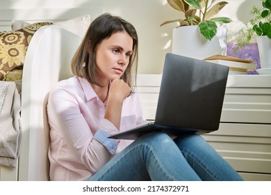 Serious sad young woman looking at laptop screen, sitting on floor at home