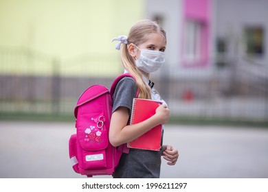 Serious Schoolgirl Wearing Medical Face Mask And Carrying A Pink Backpack Stands Near A School. Back To School. First Day At School, The Beginning Of School Year. 