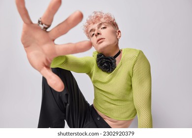 Serious self confident androgynous pink haired man keeps hand towards camera wears green net jumper trousers poses against white background. Pink haired gay looks through fingers. Homosexuality - Powered by Shutterstock