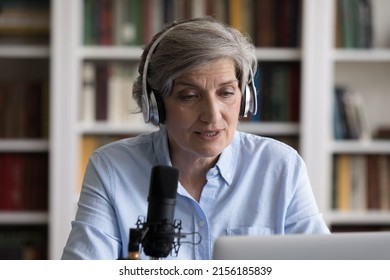 Serious senior blogger, newscaster, radio host in headphones speaking at professional microphone at laptop, holding program on air, broadcasting, recording audio book, using sound studio equipment