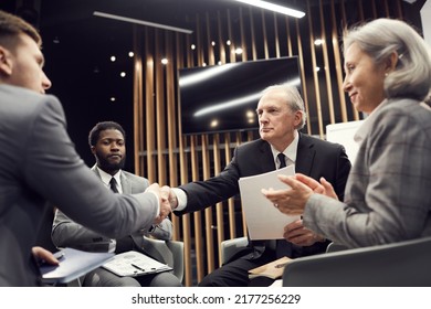 Serious Senior Businessman In Suit Sitting In Circle Of Colleagues And Handshaking With Business Group Member At Meeting