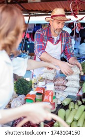 Serious Senior Caucasian Farmer Selling His Organic Vegetables On Farmers Market. Mature Woman Buying Fresh Vegetables In Outdoor Marketplace