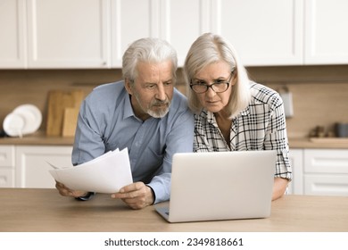 Serious senior couple checking domestic paper documents at kitchen table, using online financial banking application on laptop, doing household accounting paperwork - Powered by Shutterstock