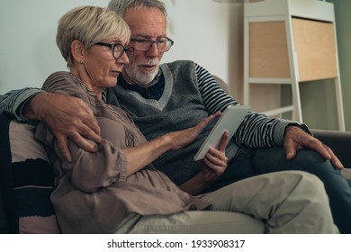 Serious Senior Couple Using Digital Tablet Together At Home. Worried Senior Adult Couple Sitting At Couch At Living Room, Talking, Typing Message And Looking In White Digital Tablet