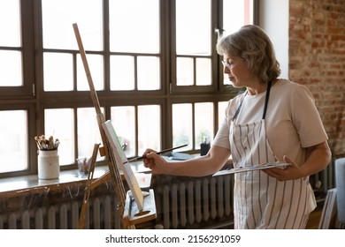 Serious Senior Female Painter Stands Near Easel Hold Palette And Paintbrush Creates Picture Engaged In Painting On Canvas, Creative Activity, Spend Retirement In Workshop, Enjoy Hobby For Soul Concept