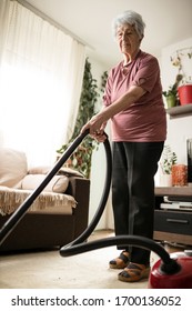 Serious Senior Gray-haired Caucasian Woman In Casual Clothes Vacuuming Carpet On The Living Room Floor. Low Angle View