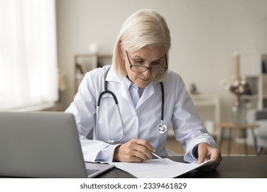 Serious senior head doctor woman working at office workplace, reading medical record, paper document at laptop, reviewing patient history, sitting at table with computer - Powered by Shutterstock