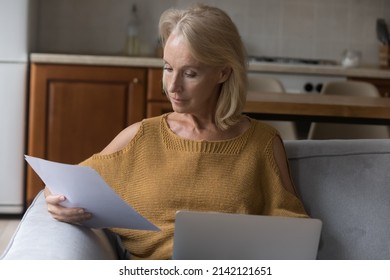 Serious Senior Homeowner Lady Doing Domestic Paperwork At Home, Reading Paper Document At Laptop, Paying Paper Bills, Invoices, Insurance Fees, Reading Agreement, Letter, Notice, Sitting In Couch