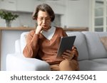 Serious senior lady using digital tablet, sitting on sofa with device, reading online news or learning new application. Older gen and modern tech