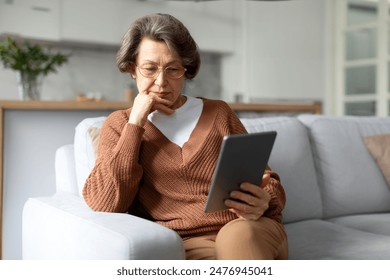 Serious senior lady using digital tablet, sitting on sofa with device, reading online news or learning new application. Older gen and modern tech - Powered by Shutterstock