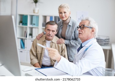 Serious Senior Male Doctor In Eyeglasses Sitting At Desk And Using Computer While Showing Test Results To Man Supported By Wife At Consultation