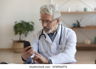 Serious Senior Male Doctor Look At Cellphone Screen Talk Speak On Video Call With Patient Online. Focused Mature Man GP Have Webcam Digital Consultation With Client In Hospital. Virtual Event Concept.