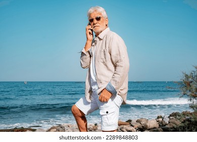 Serious Senior Man at the Beach Using Phone - Vacation Retirement Lifestyle at Sea - Angry Elderly Grandfather Talking on Mobile Phone Standing close to the Sea Water - Powered by Shutterstock