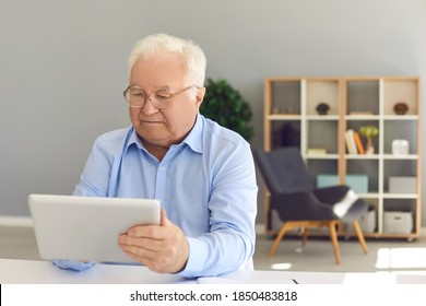 Serious Senior Man Sitting At Desk At Home With Tablet Computer In Hand, Booking Medical Appointment Online, Using Telemedicine Service, Checking Pension Plan Website Or Reading News On Social Media