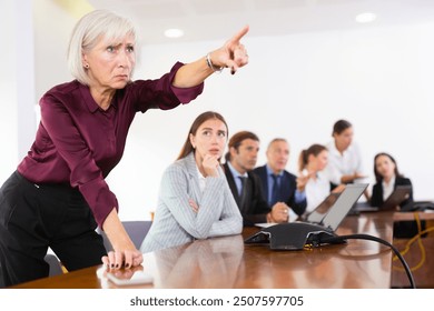 Serious senior woman entrepreneur leading daily briefing in meeting room in company office, standing at desk and making pointing finger gesture. - Powered by Shutterstock