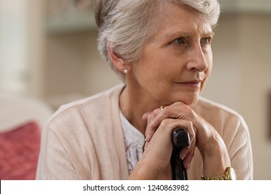 Serious Senior Woman Sitting On Couch Holding Walking Stick And Looking Away. Portrait Of Thoughtful Old Grandmother Leaning On Cane. Elderly Sad Lady With Her Walking Stick.