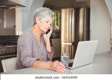 Serious Senior Woman Talking On Mobile Phone While Sitting In Kitchen