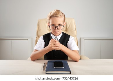 Serious Small Entrepreneur Sitting At Big Table And Looking At Camera